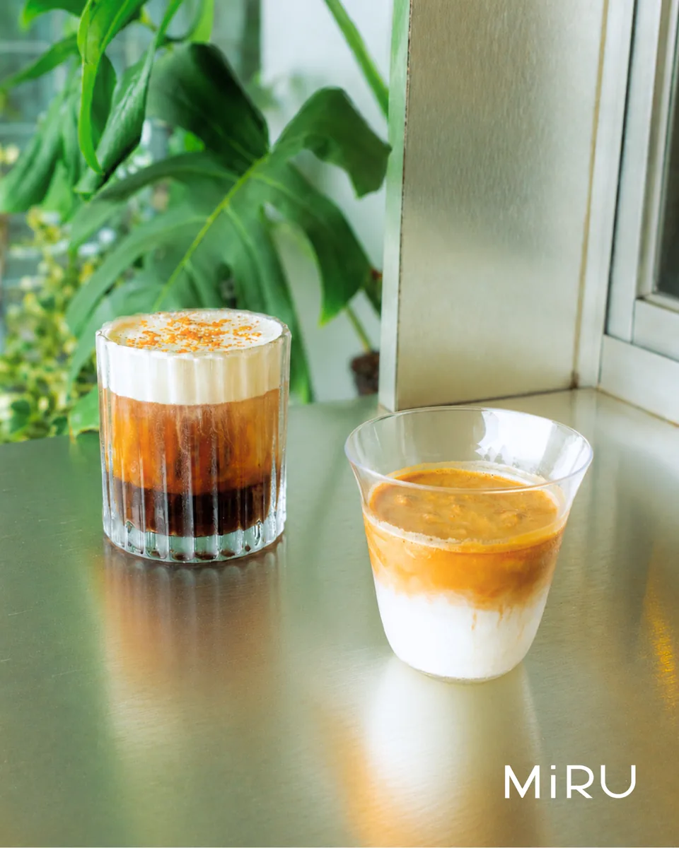 Dua minuman MiRU di dekat jendela: es kopi berlapis dengan zest jeruk dan minuman krimi berwarna oranye