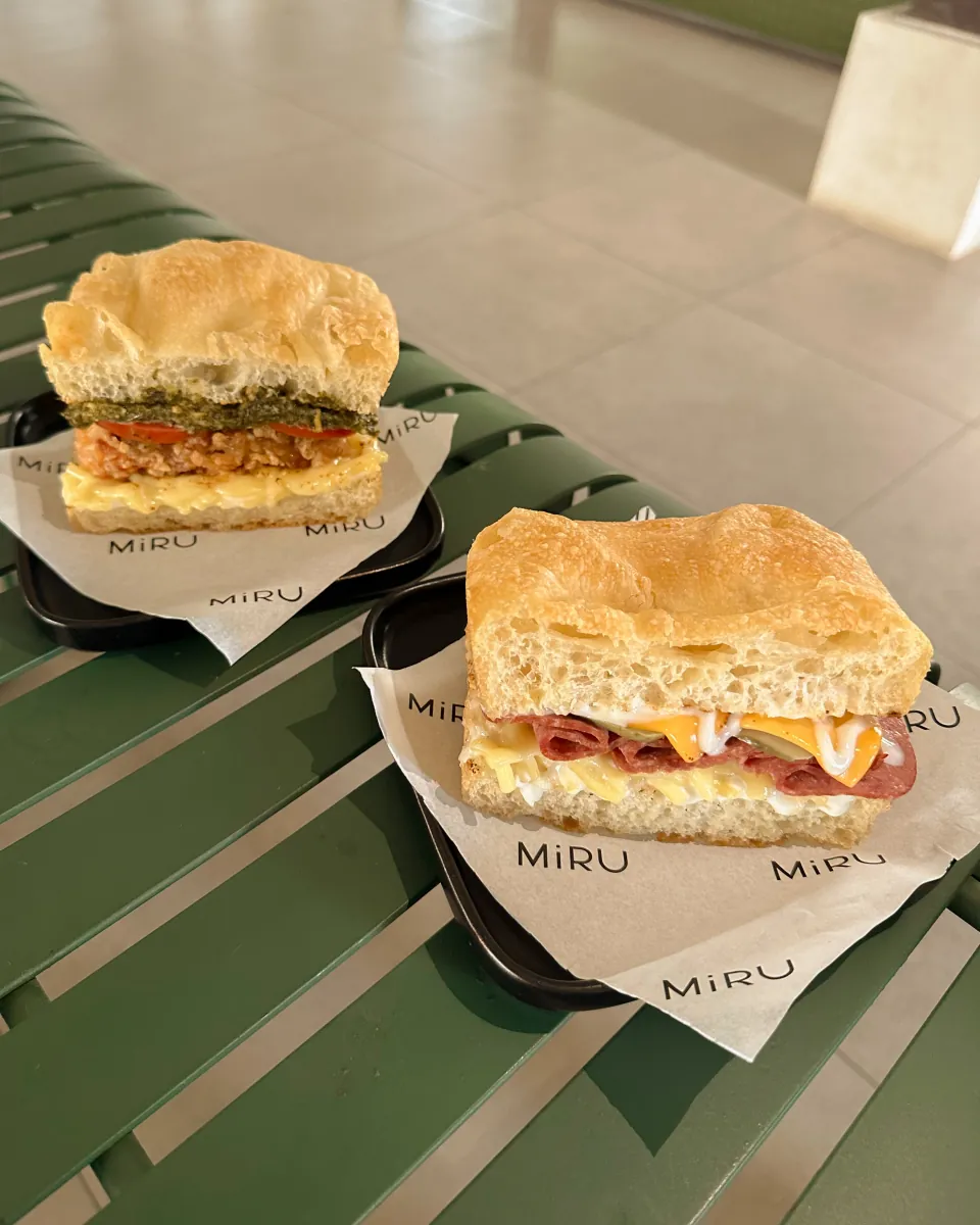 Dua sandwich focaccia MiRU di atas kertas cetak MiRU di bangku hijau